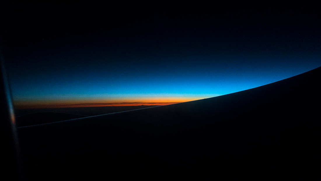 A sunset from a plane, the black ground being topped by an orange, a yellow and then a blue band, before the sky returns to black on top of the picture.