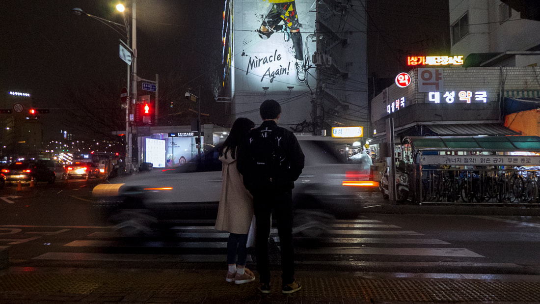 Two lovers in Seoul waiting by the side of the road at night before a graffiti which says : 