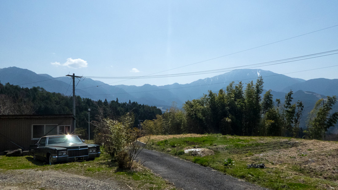 Car parked in the sun surrounded by grenery, with a chain of mountain on the back. Magome, Japan.